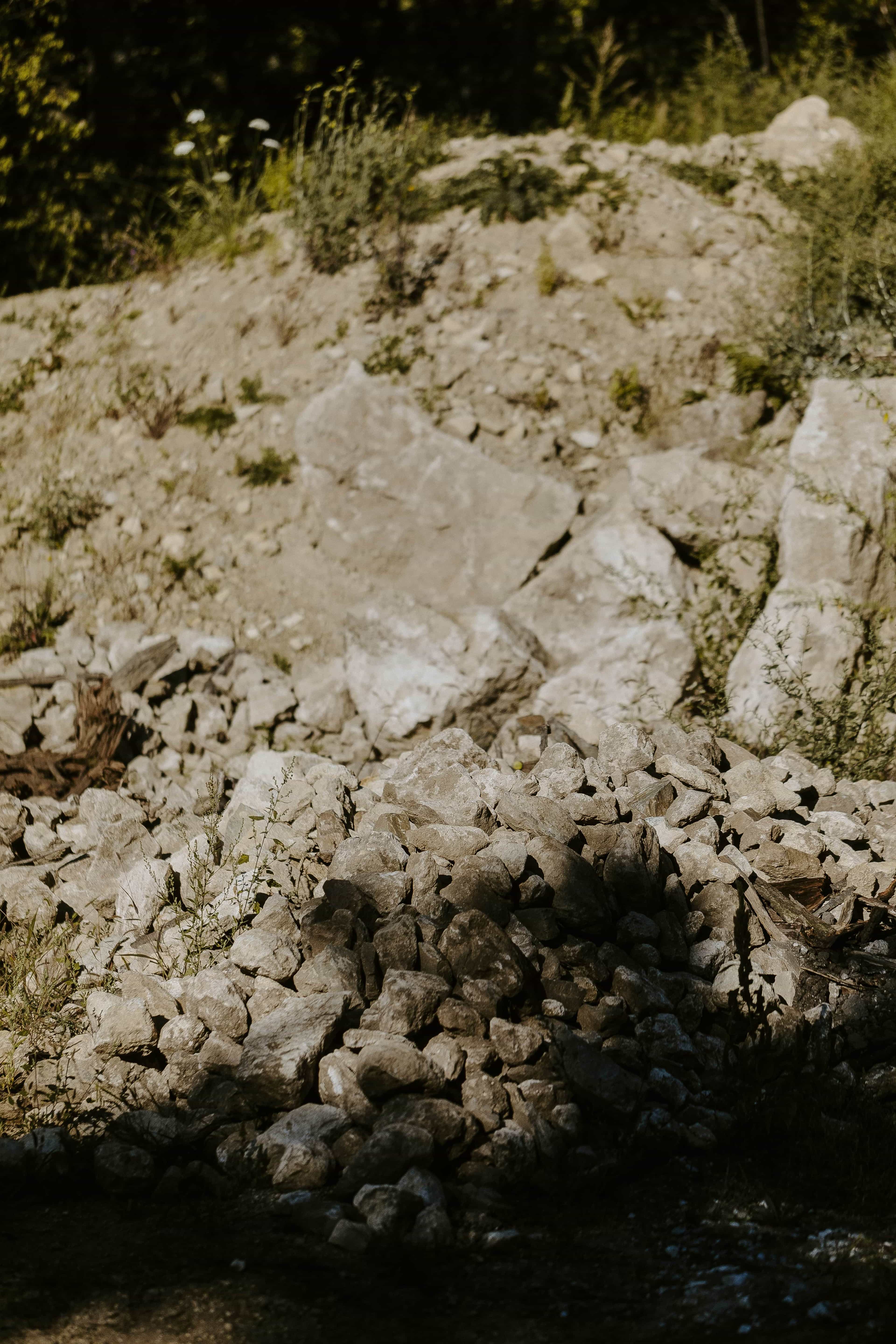 A pile of rocks and stones in front of a rocky hillside with patches of greenery.