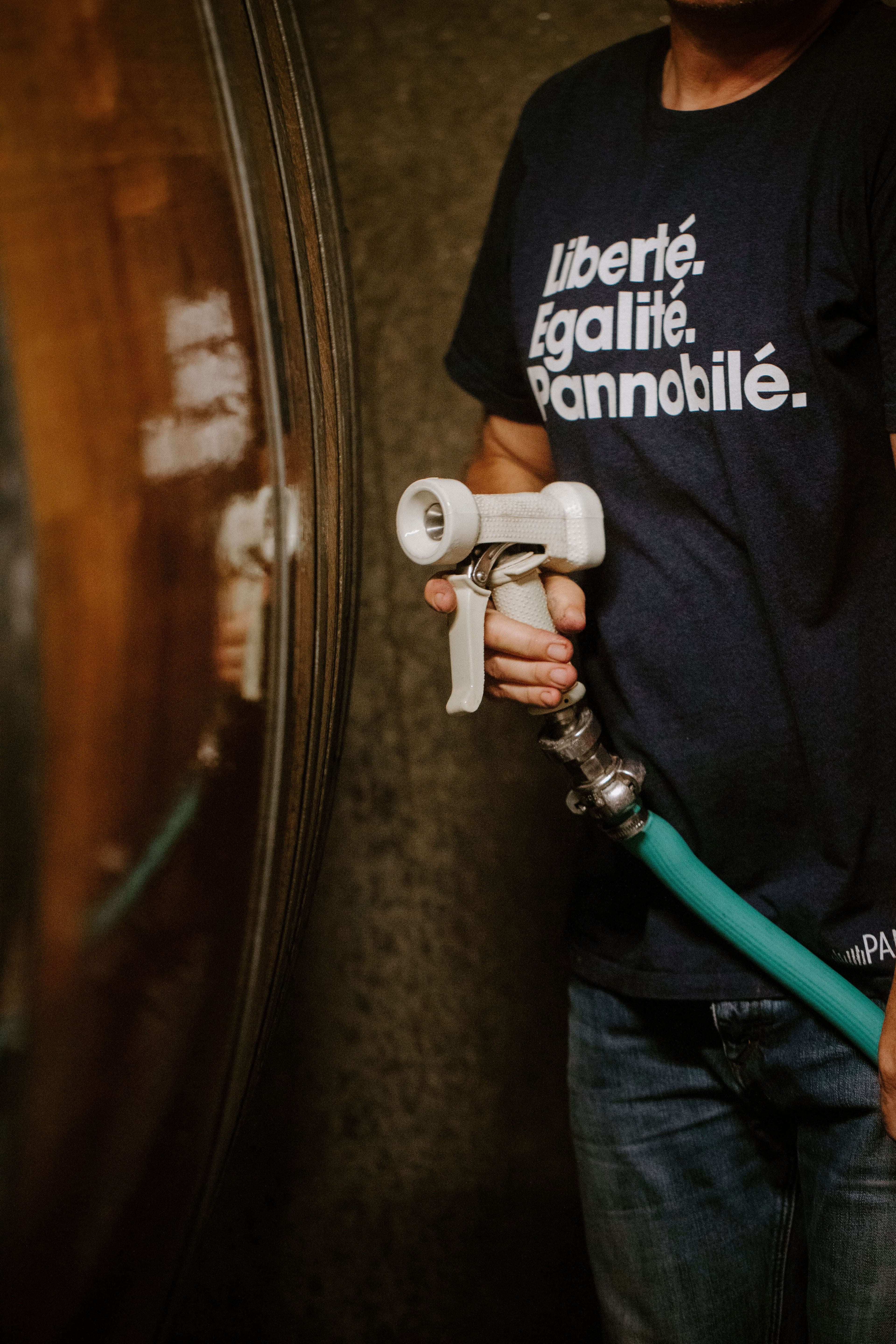 A person in a dark shirt with text "Liberté, Égalité, Pannobile" holds a spray nozzle attached to a green hose. The scene is industrial, with metal tanks.