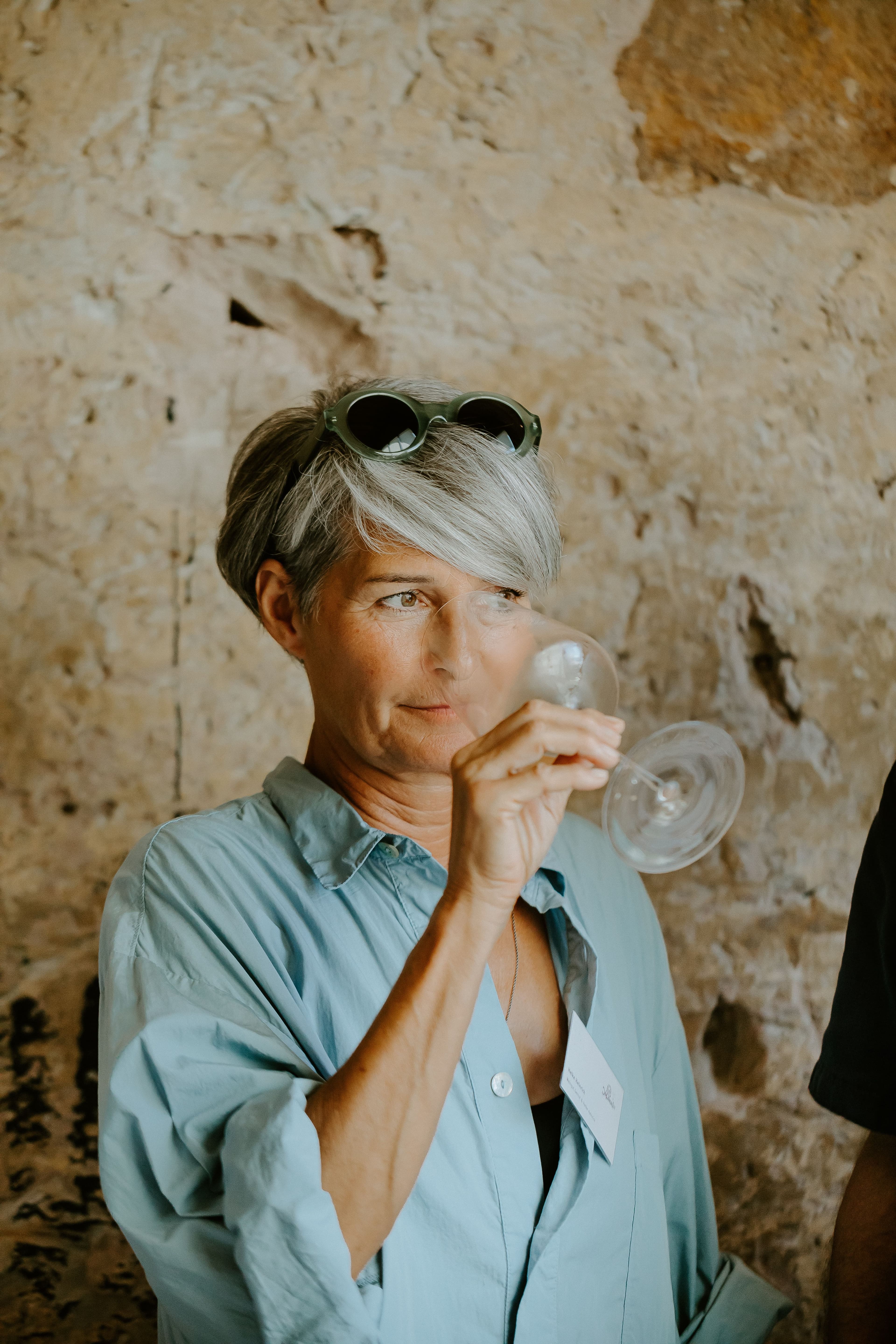A person with short gray hair, wearing sunglasses on their head and a light blue shirt, holds a wine glass in a rustic setting, conveying curiosity.