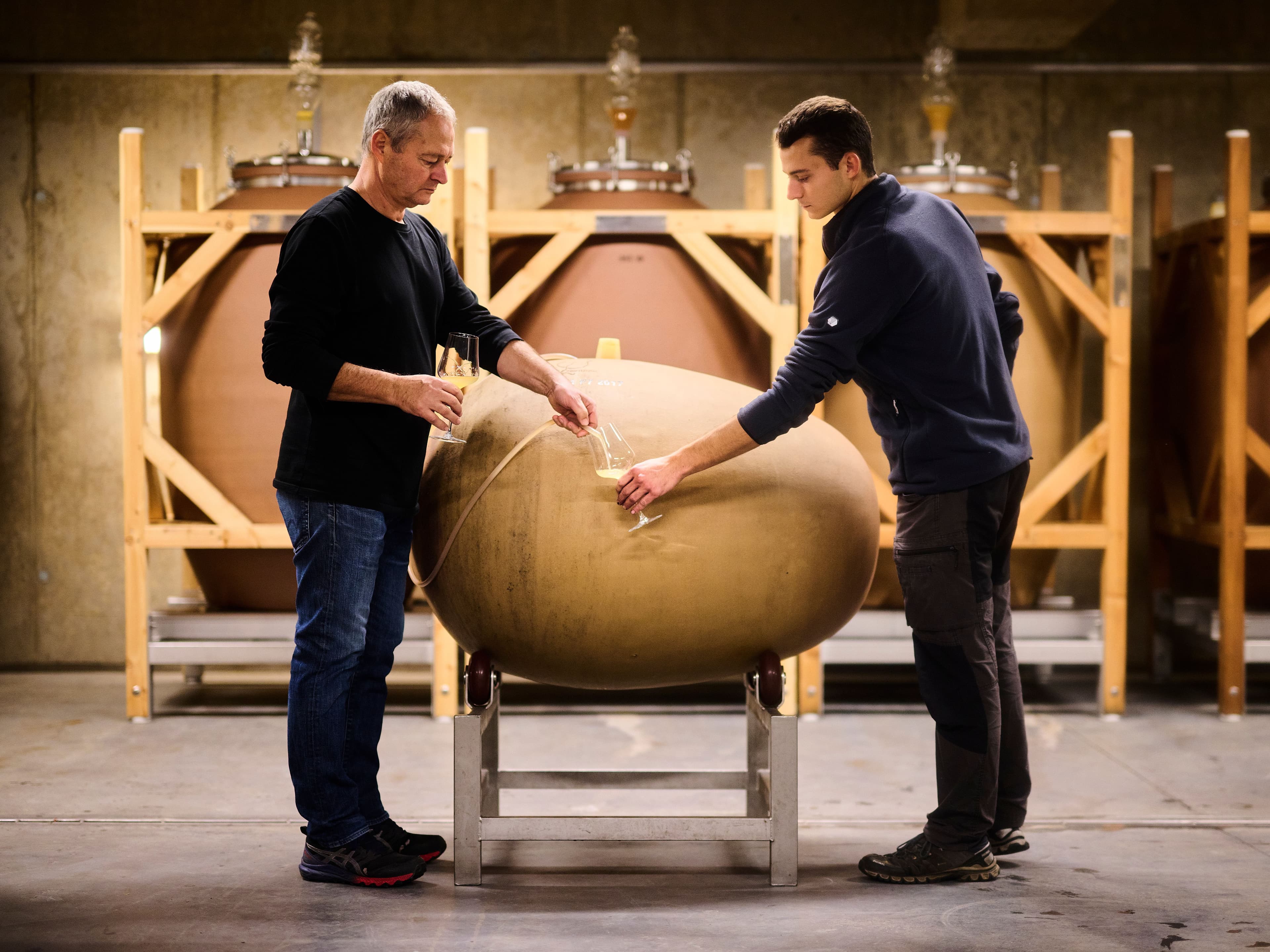 Two men carefully inspect a wine barrel in a dimly lit cellar, surrounded by large clay fermentation vessels. The scene conveys concentration and expertise.