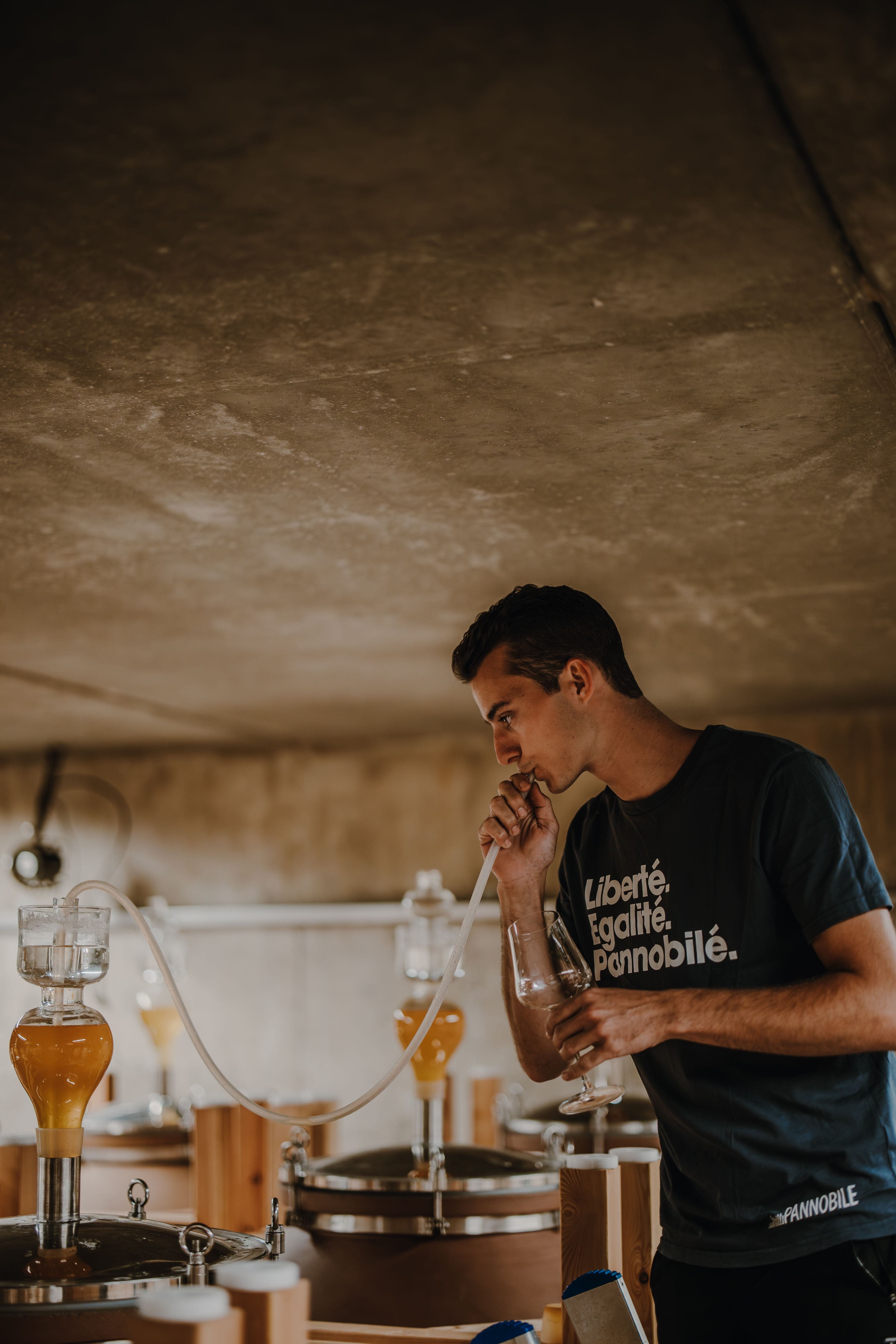 Man in a dark t-shirt samples a drink using a tube connected to a glass container with amber liquid. The setting is a dimly lit, rustic room.