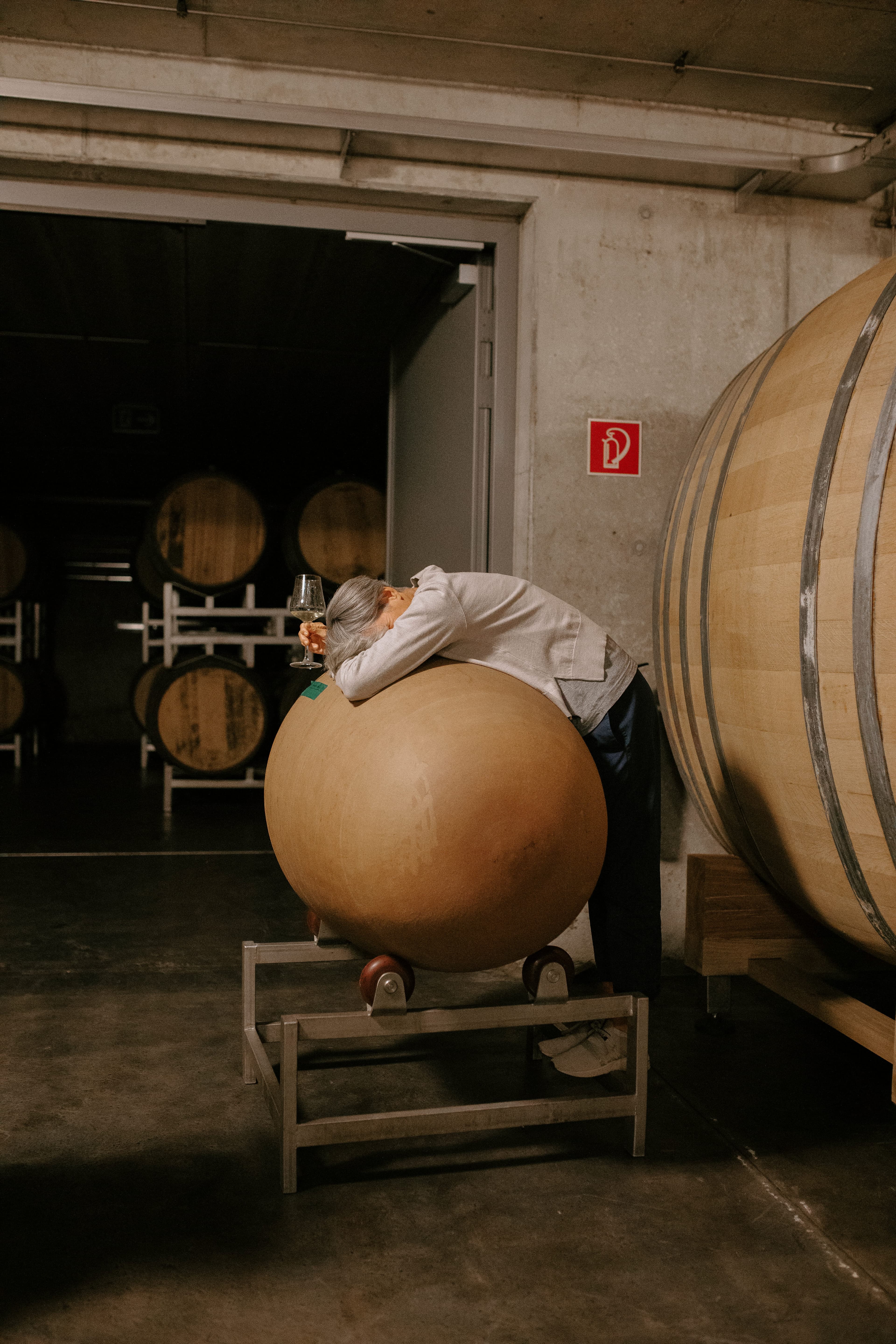 A person leans over a large round vat in a dimly lit wine cellar, holding a glass of wine. Wooden barrels and a fire extinguisher are visible in the background.