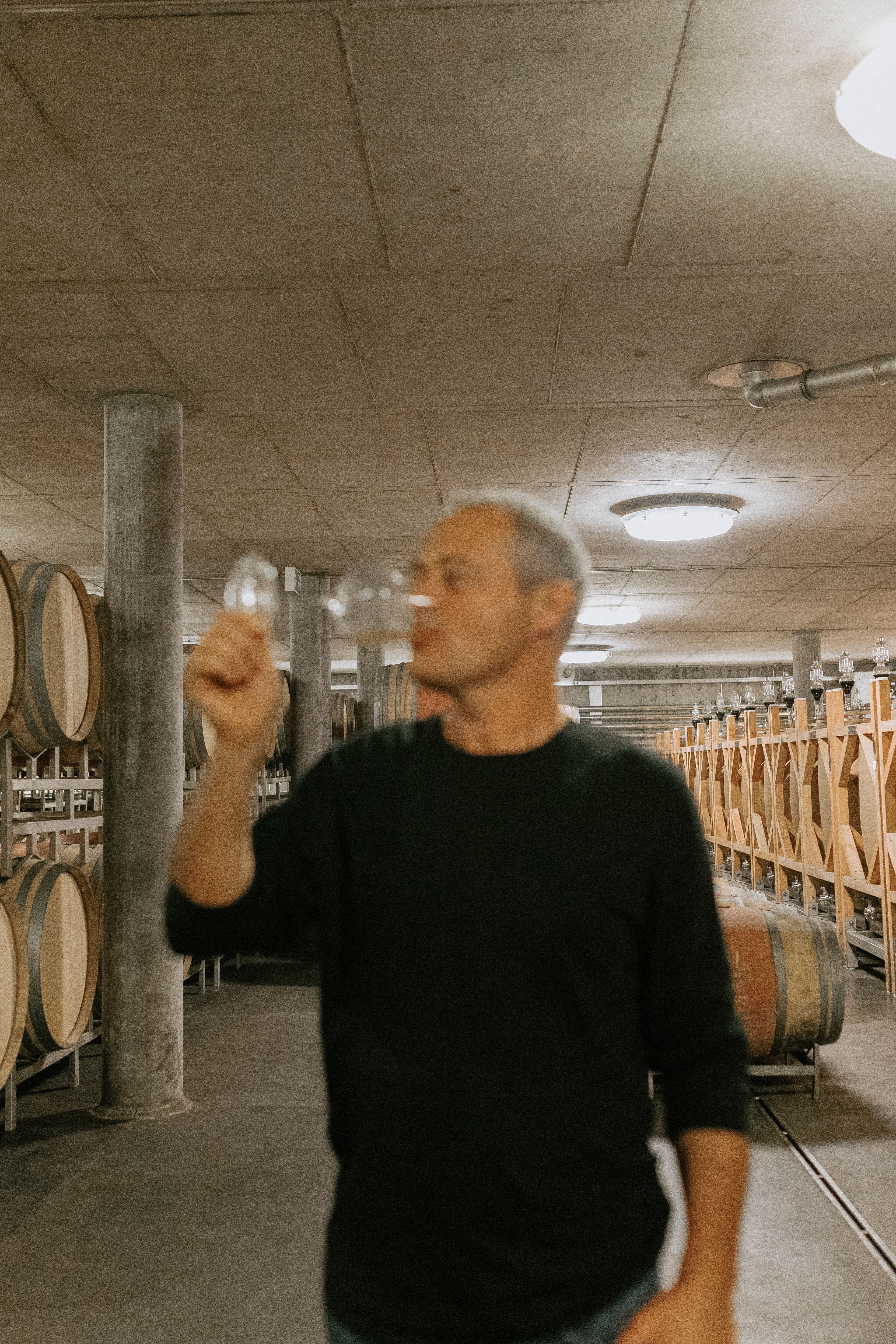 A person in a dimly lit wine cellar tastes wine from a glass; rows of wooden barrels line the walls, creating a focused, contemplative atmosphere.