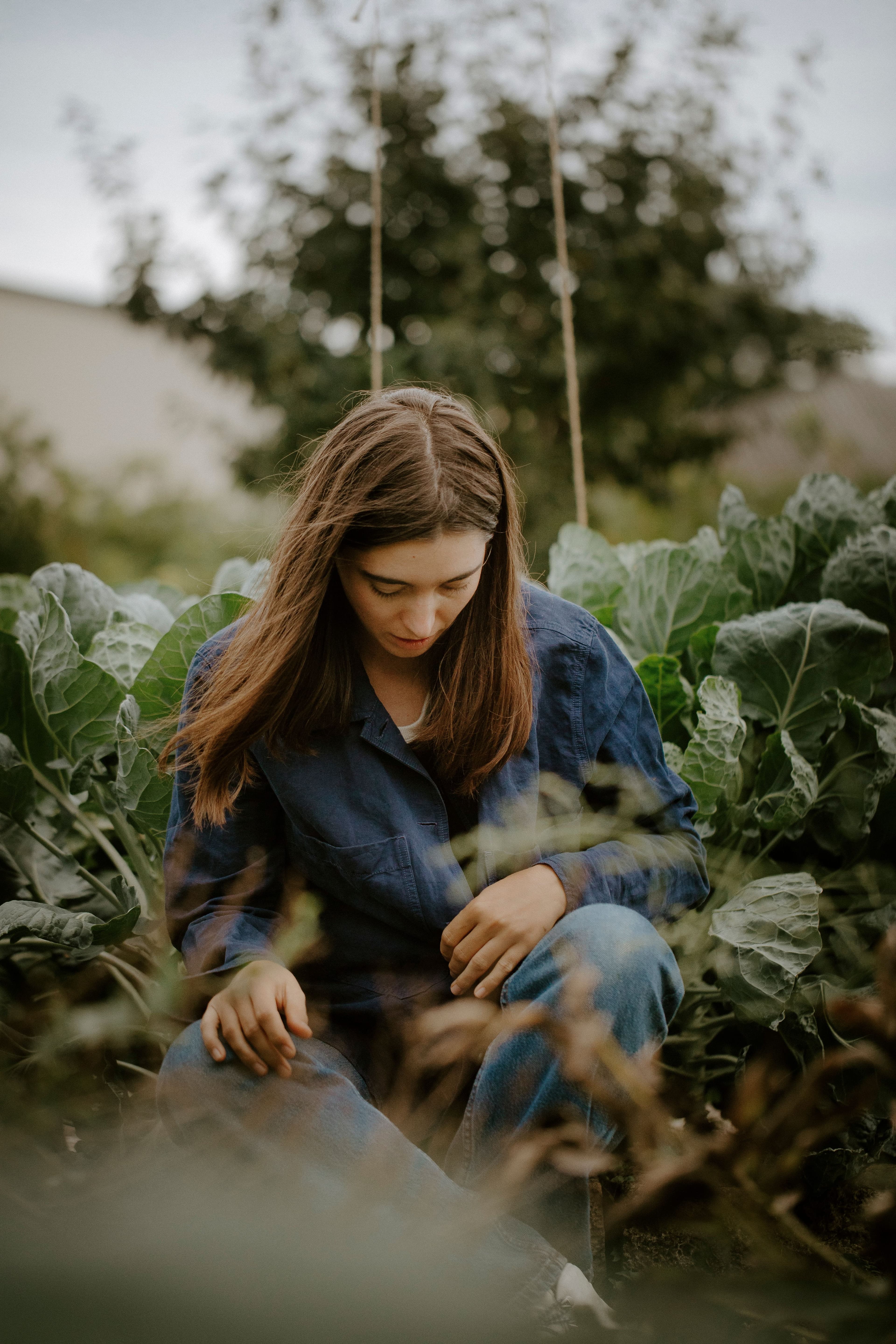 A woman with long brown hair, wearing a blue outfit, kneels thoughtfully among lush green plants in a serene garden setting, under an overcast sky.